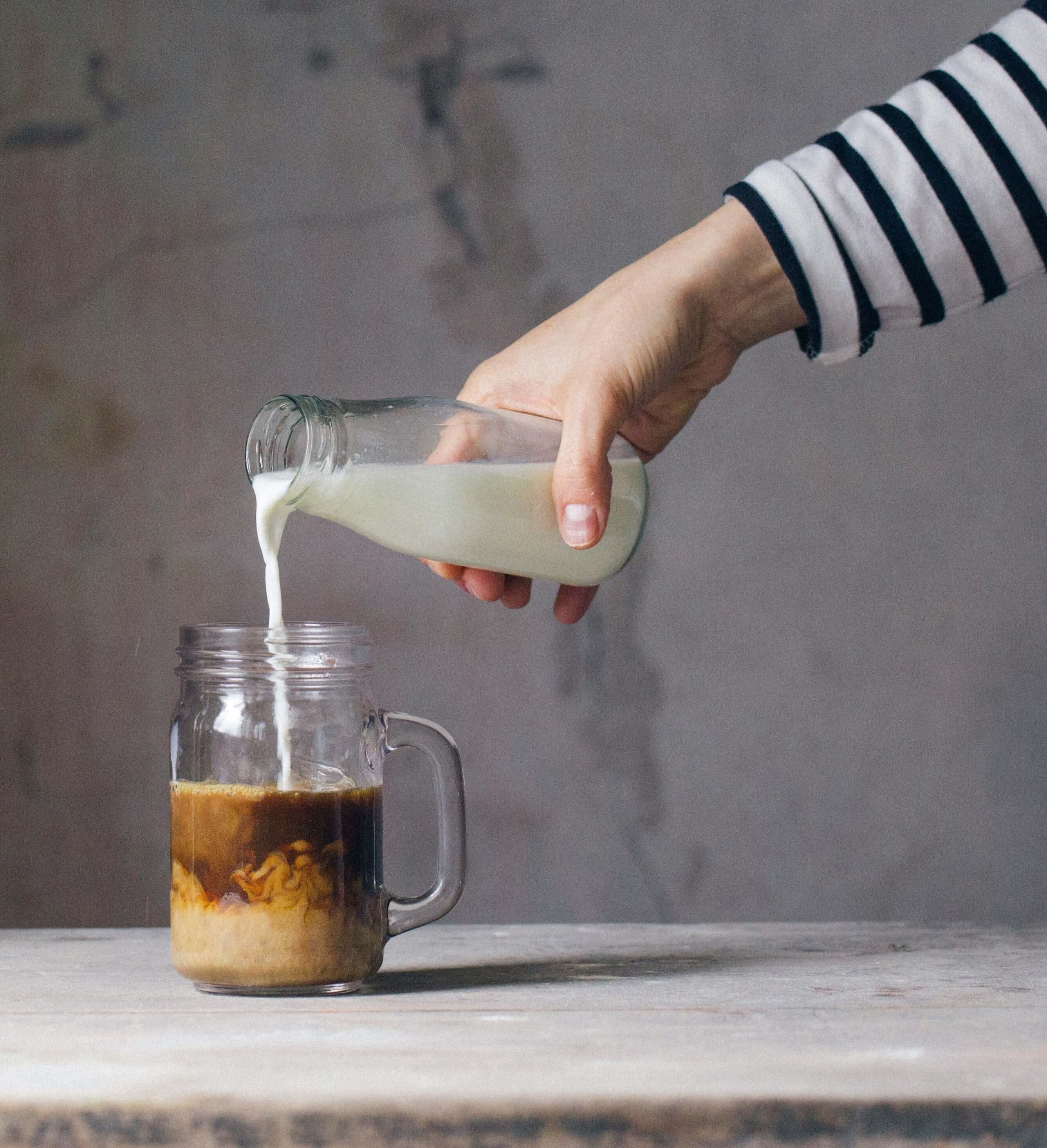 hand pouring carafe of milk into a jar of coffee placed on a table.